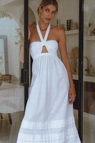 Woman wearing a white linen maxi dress with halter neck and front cut out standing in a room with decor elements.