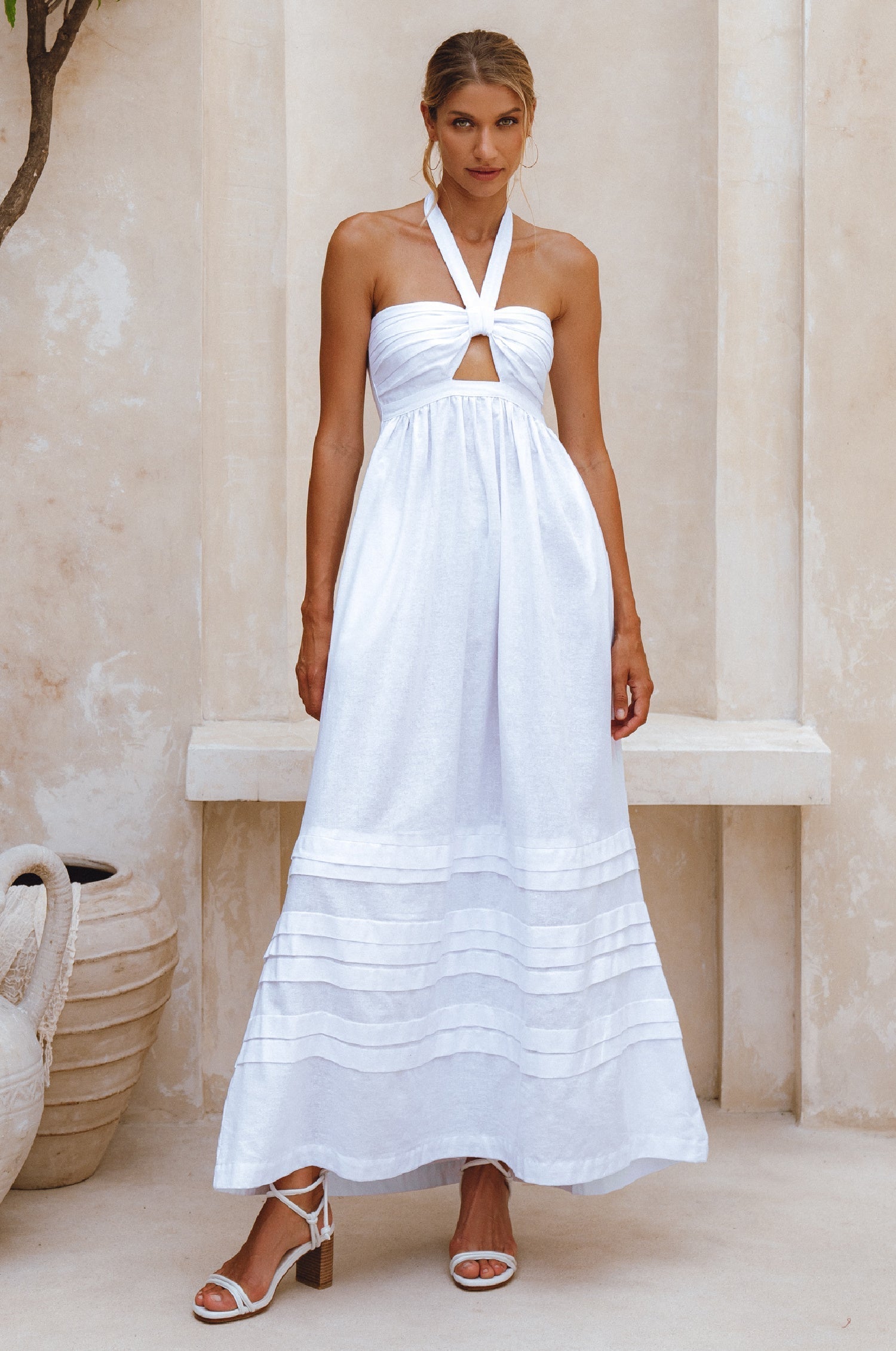 Woman wearing a white linen maxi dress with halter neck and front cut out standing in a sunlit room with columns.