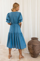 Woman wearing a blue dress standing next to a large vase against a white curtain background back view