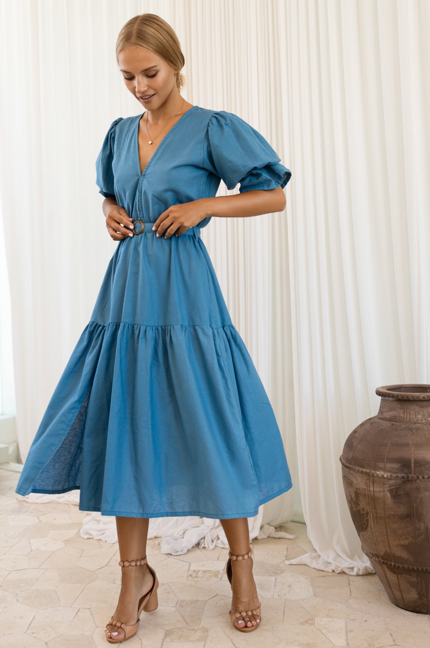Woman wearing a blue dress standing in a room with white curtains and a large vase.