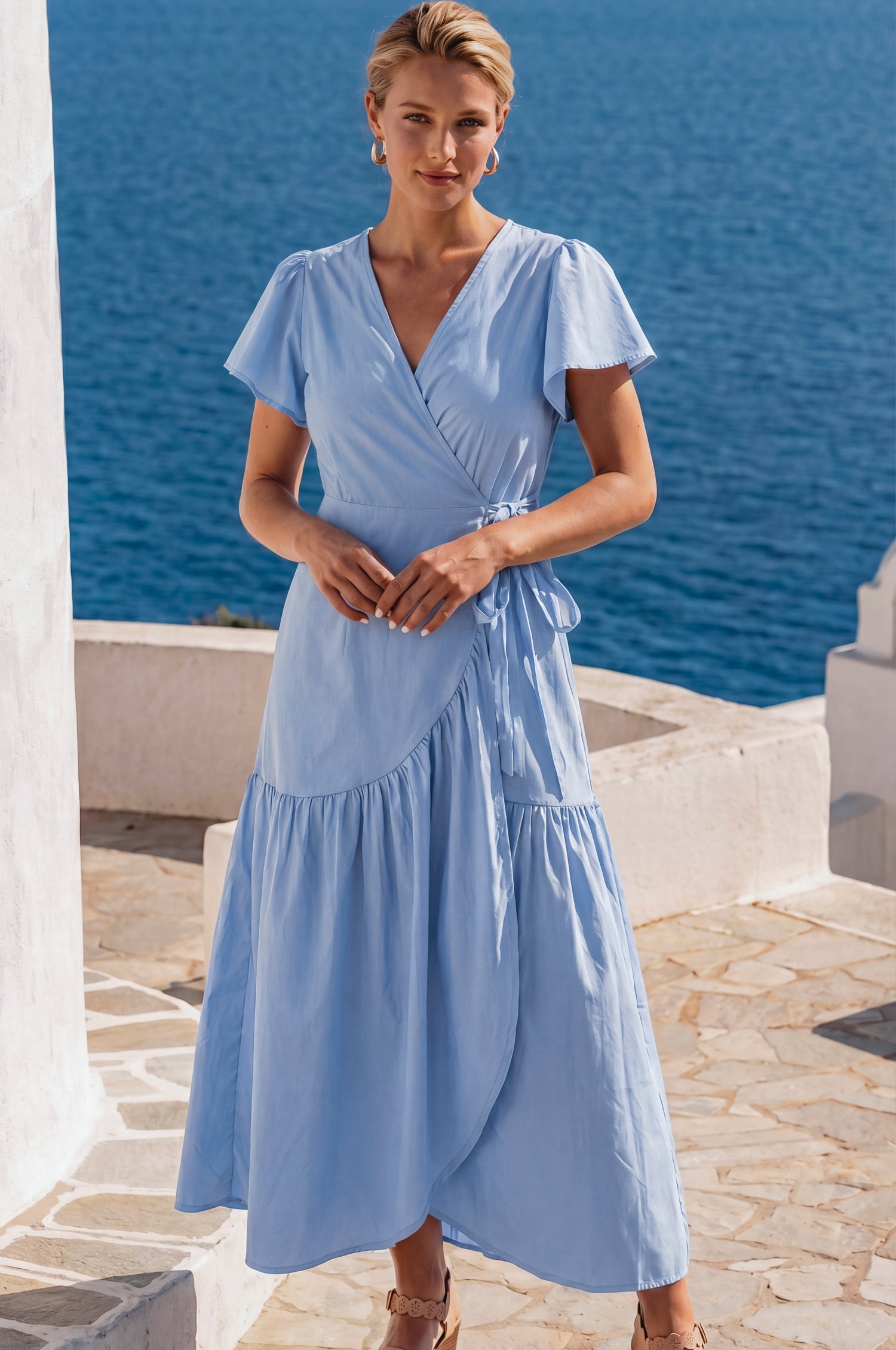 Woman in a light blue wrap style midi dress with flutter sleeves and v-neckline standing by a scenic waterfront, front view.