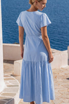 Woman wearing a light blue dress flutter sleeves standing by a body of water, back view.