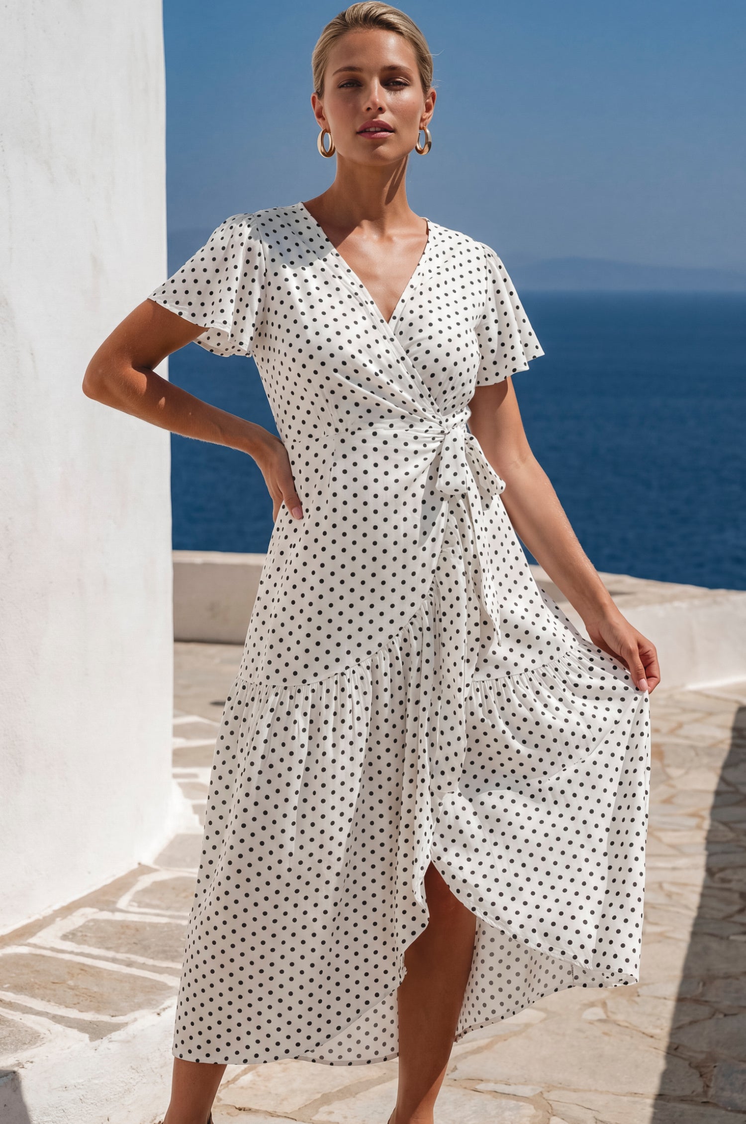 Woman wearing a white wrap-style midi dress with black polka dots, v-neckline and flutter sleeves by the ocean.
