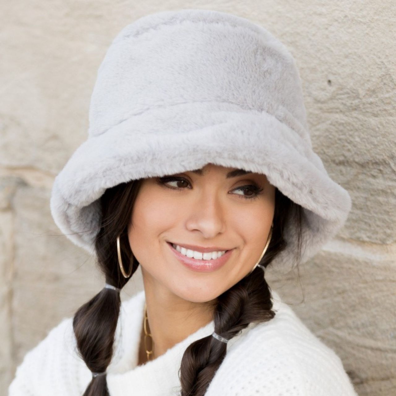 Woman wearing a gray plush faux fur bucket hat, holding the brim, side view against a stone wall