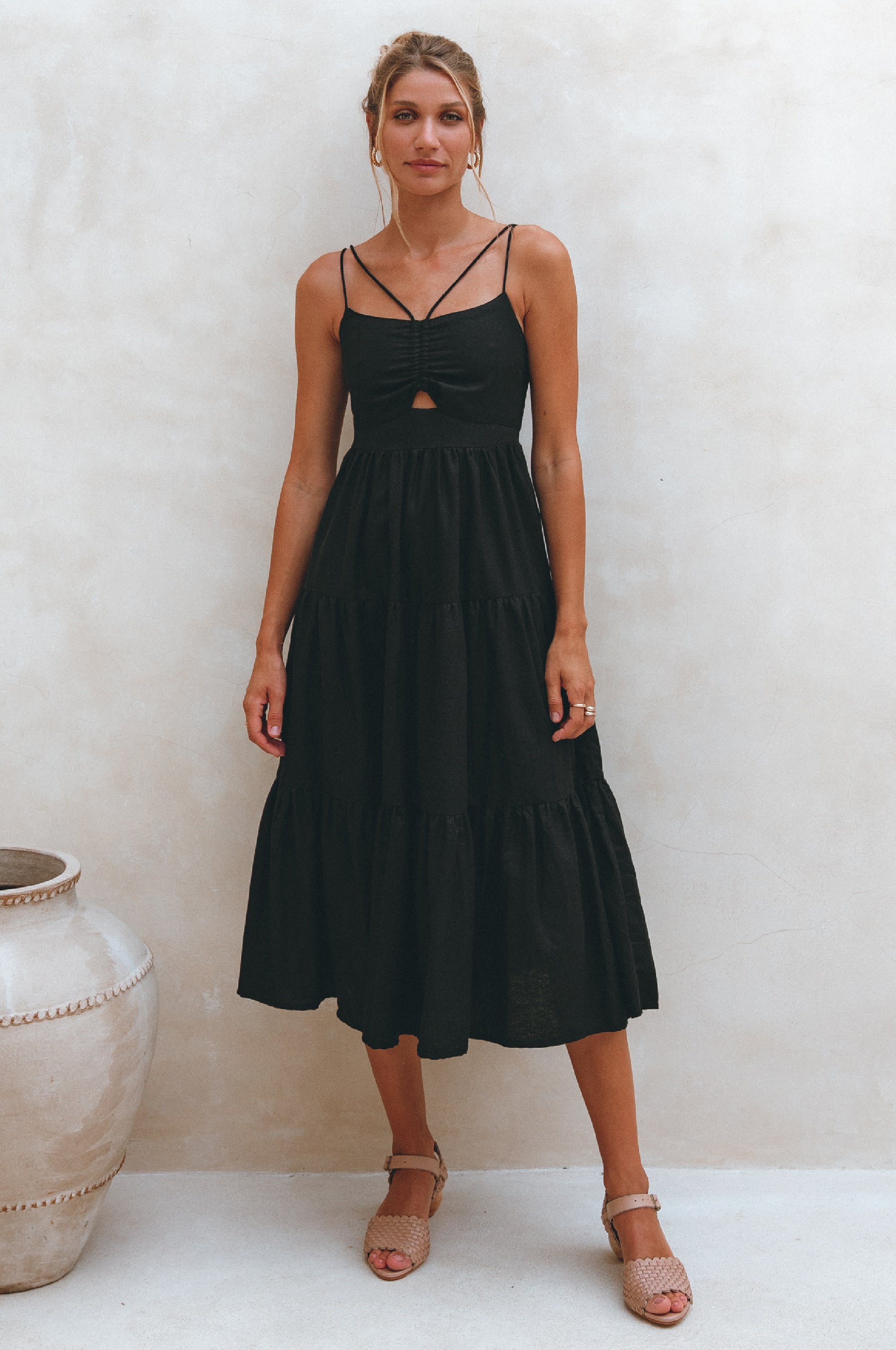 Woman wearing a black linen tiered midi dress with dual adjustable spaghetti straps standing against a white wall with a decorative vase.