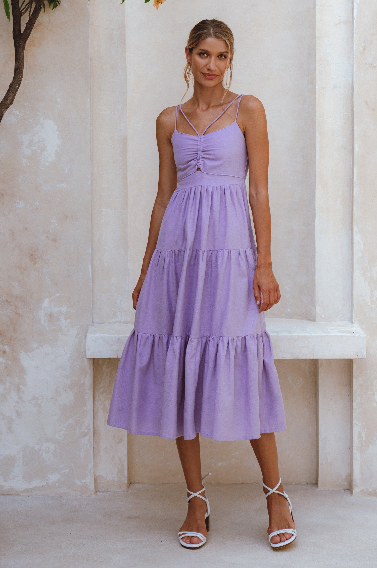 Woman wearing a lavender linen tiered midi dress with dual adjustable spaghetti straps standing against a light-colored wall, front view.
