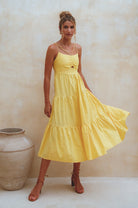 Woman wearing a yellow dress linen tiered midi dress with dual adjustable spaghetti straps standing against a beige wall with a decorative vase.