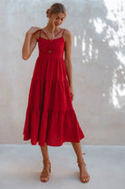 Woman wearing a red linen tiered midi dress with dual adjustable spaghetti straps standing against a light-colored wall