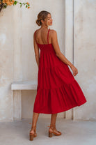 Model wearing a red linen tiered midi dress with dual adjustable spaghetti straps standing against a light-colored wall, back view