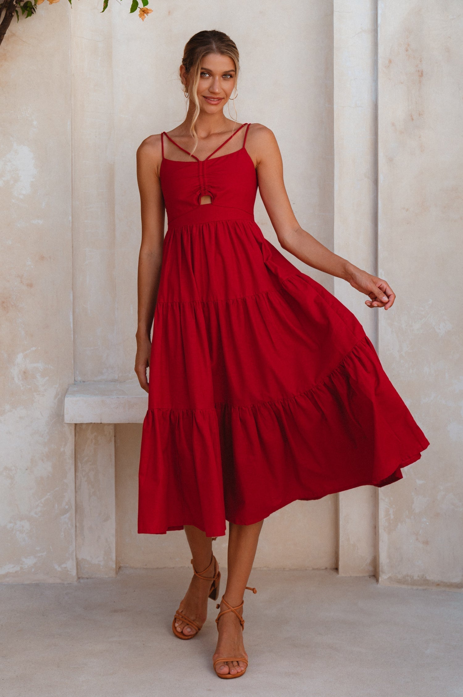 Woman wearing a red linen tiered midi dress with dual adjustable spaghetti straps standing against a light-colored wall, front view.