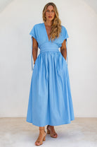 Woman wearing a light blue maxi dress standing with hands in pockets against a white background