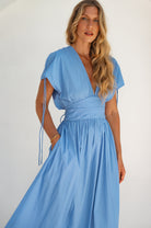 Woman wearing a blue maxi dress with sleeves un-gathered, deep v-neckline and hand in pocket against a white background