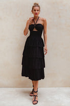 Woman wearing a black halter neck, shirred bodice, tiered midi dress against a beige background