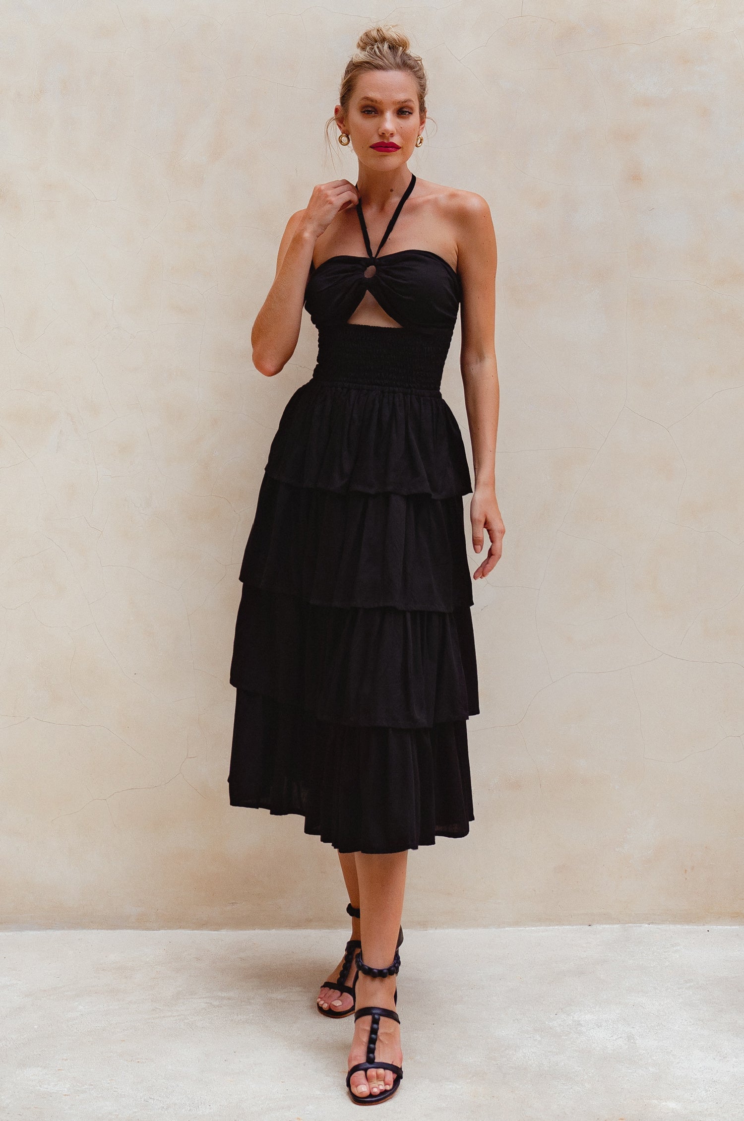 Woman wearing a black halter neck, shirred bodice, tiered midi dress against a beige background