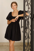 Woman wearing a black mini dress with puff sleeves standing next to an ornate metal gate.