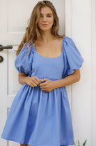 Woman wearing a blue mini dress with puff sleeves standing against a white door.