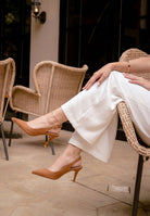 Person wearing brown high-heeled shoes and white pants sitting on a wicker chair.