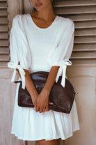 A woman wearing a white puff-sleeve mini dress with tie details on the sleeves, holding a dark brown leather clutch bag against a rustic wooden shutter background.