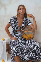 Woman in a black and white patterned dress with flutter sleeves and deep v-neckline sitting on a wood bench holding a basket of lemons against a neutral background