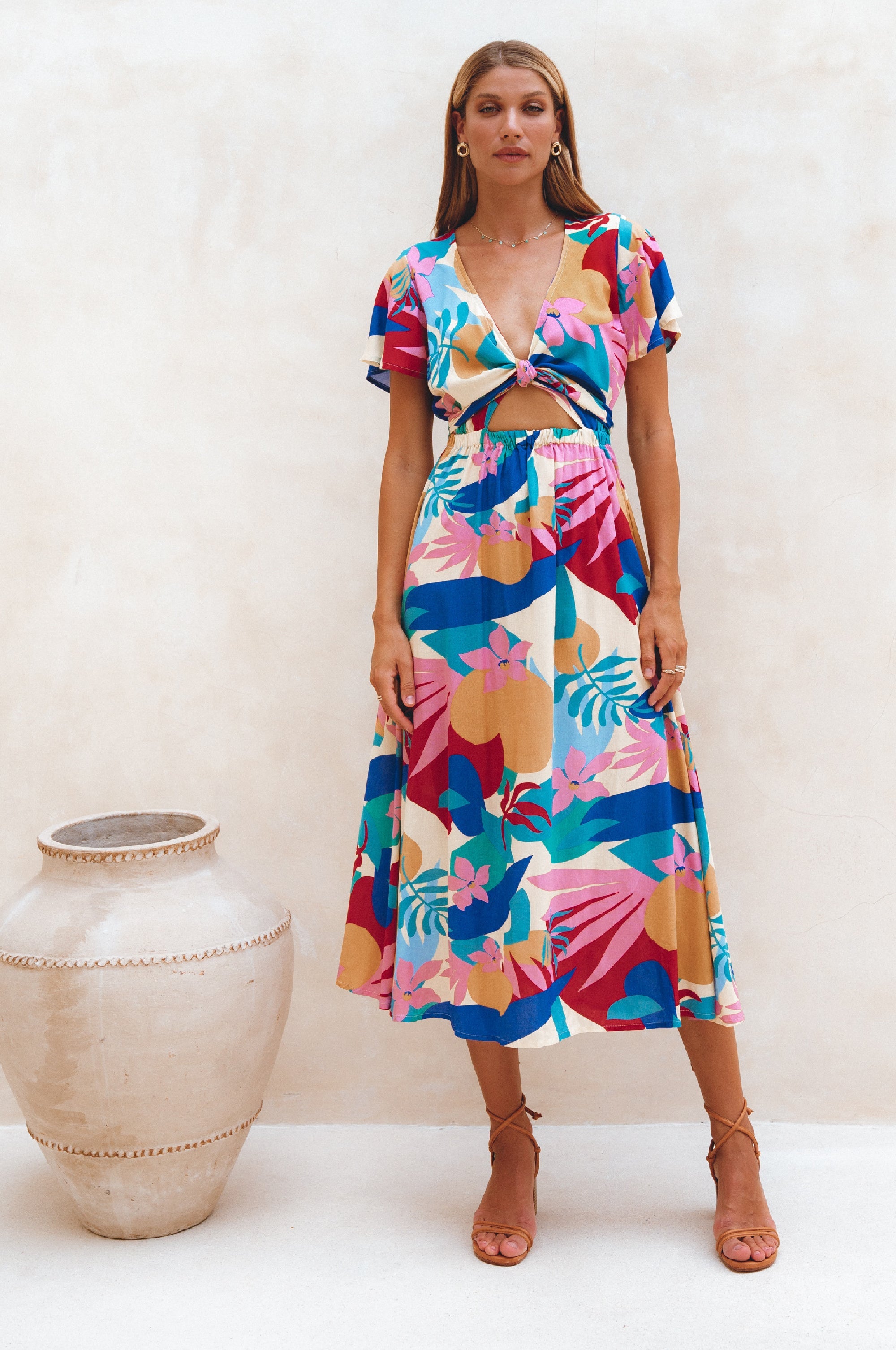 Woman wearing a colorful floral midi dress with flutter sleeves, deep v-neckline and front cut out standing next to a decorative vase on a white background
