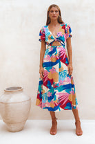 Woman wearing a colorful floral midi dress with flutter sleeves, deep v-neckline and front cut out standing next to a decorative vase on a white background