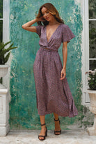 Woman in a purple floral patterned midi dress with deep v-neckline and flutter sleeves standing against a textured green wall.