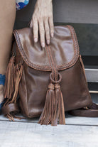 Brown leather backpack with tassels held by a hand on a wooden surface.