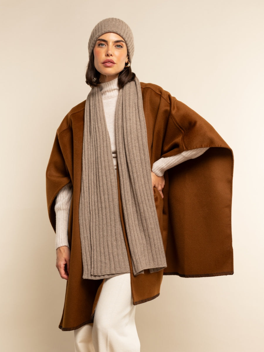 Woman wearing a brown coat, brown scarf, and knit hat against a plain background