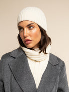 Woman wearing a cream cashmere knit beanie and scarf with a gray coat on a beige background
