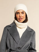 Woman wearing a cream cashmere knit beanie and scarf with a gray coat on a beige background
