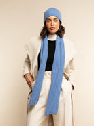 Person wearing a light blue cashmere knit scarf and beanie with a white coat on a beige background