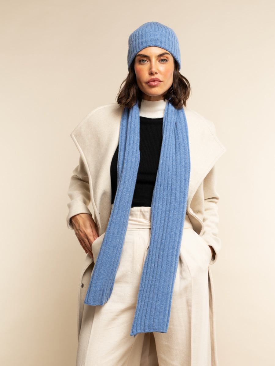Person wearing a light blue cashmere knit scarf and beanie with a white coat on a beige background