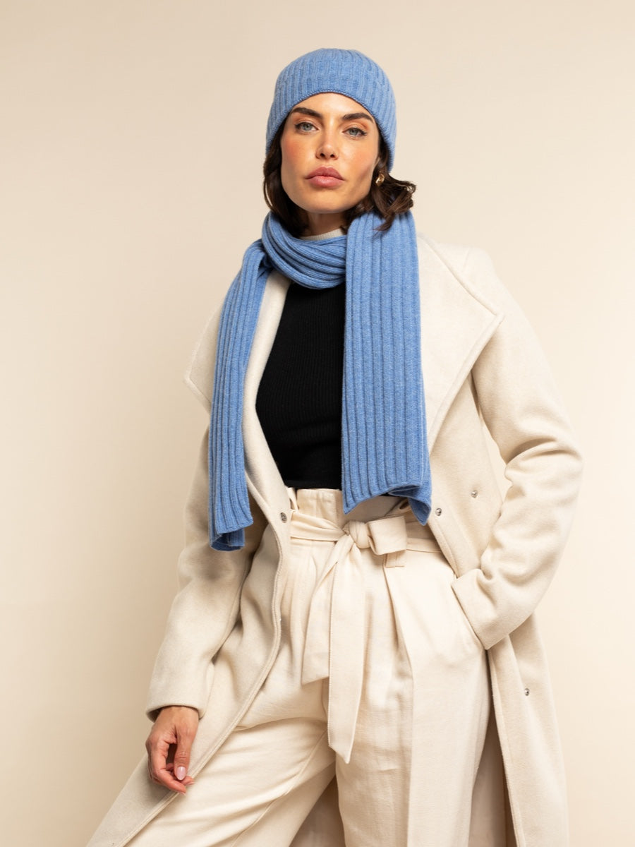 Woman wearing a light blue cashmere knit hat and scarf with a beige coat against a plain background