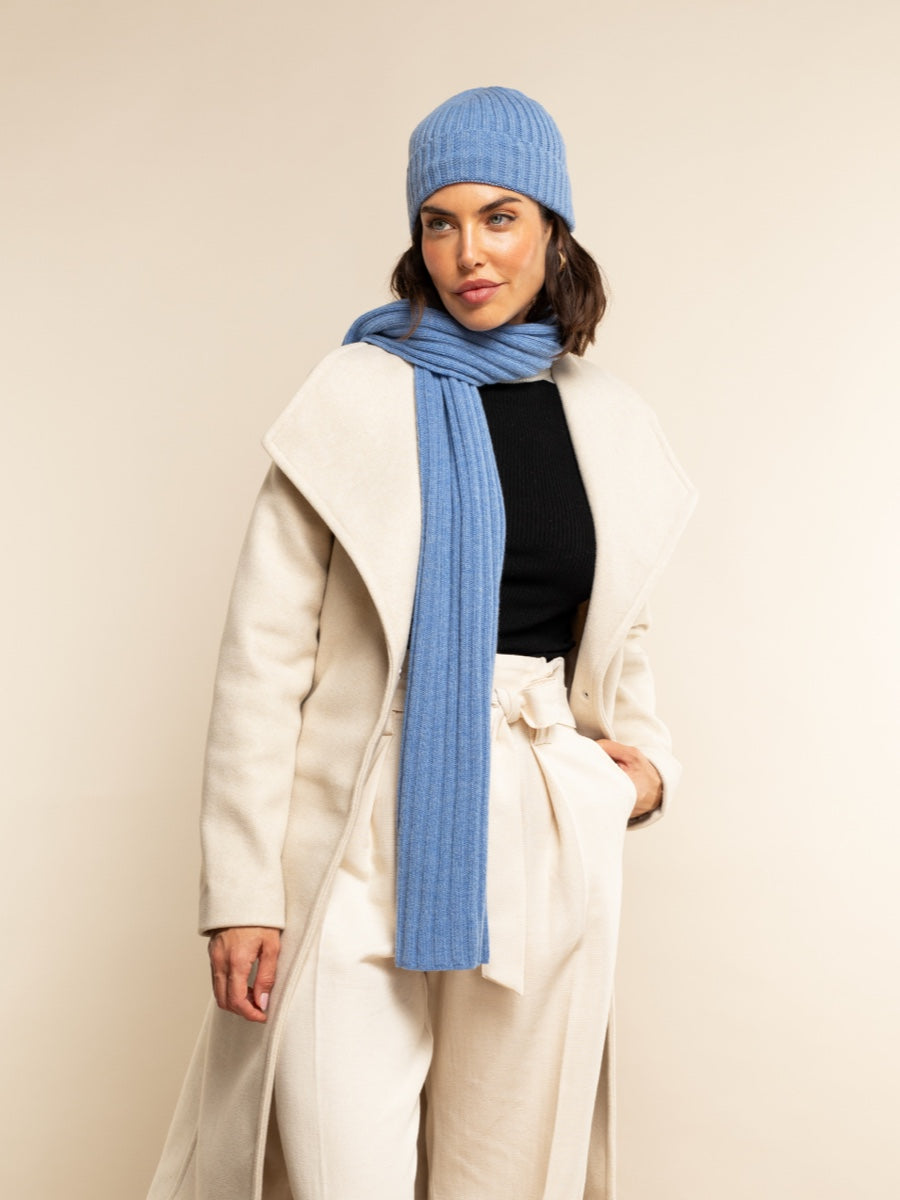 Person wearing a blue cashmere knit hat and scarf with a white coat against a beige background