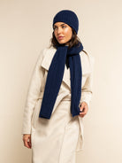 Woman wearing a white coat, navy blue cashmere beanie, and scarf against a beige background
