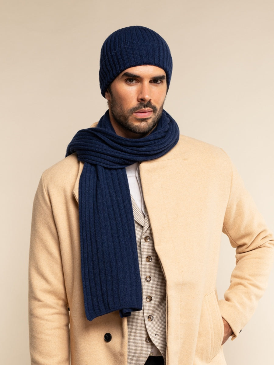Man wearing a navy cashmere knit beanie and scarf with a beige coat against a beige background