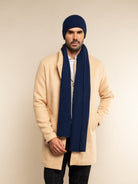 Man wearing a beige coat, cashmere navy beanie, and scarf against a beige background