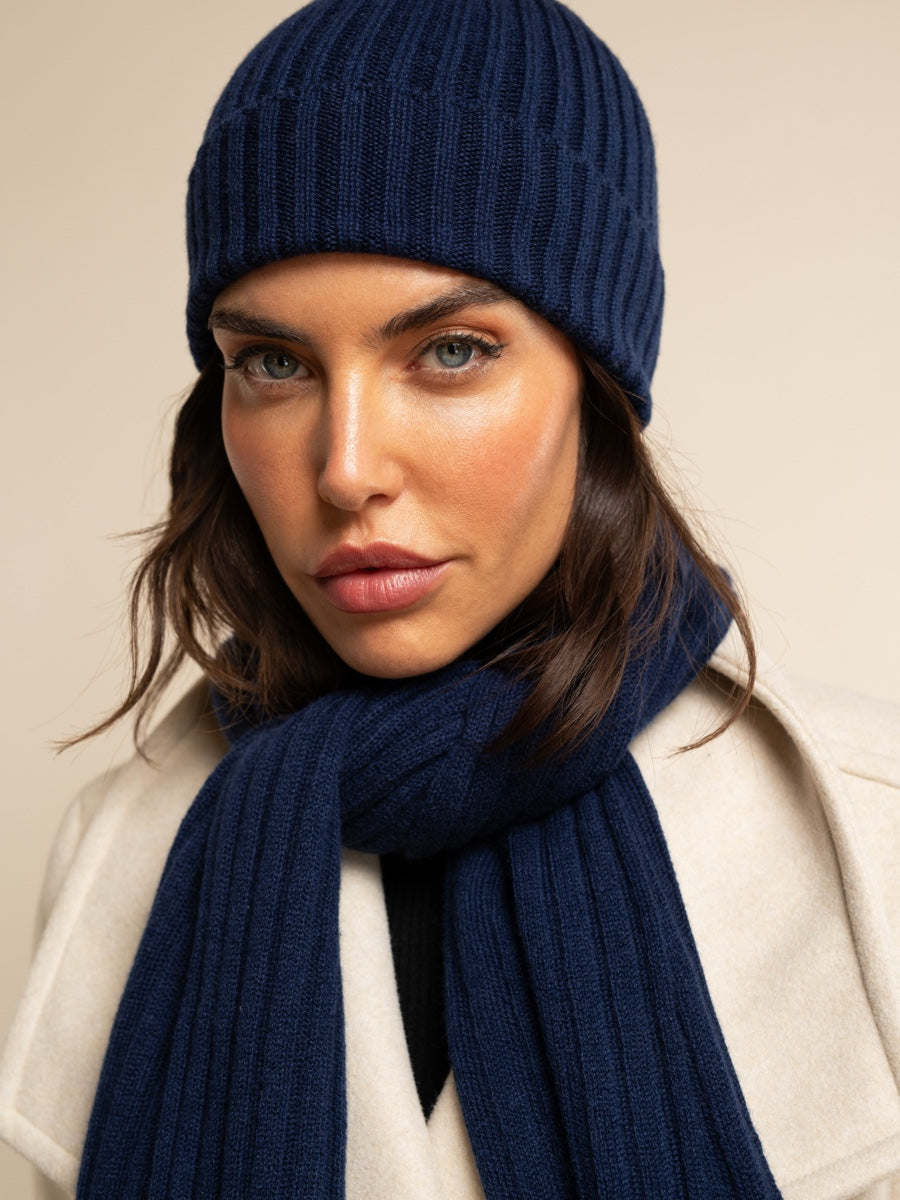 Woman wearing a navy blue cashmere   knit hat and scarf against a beige background