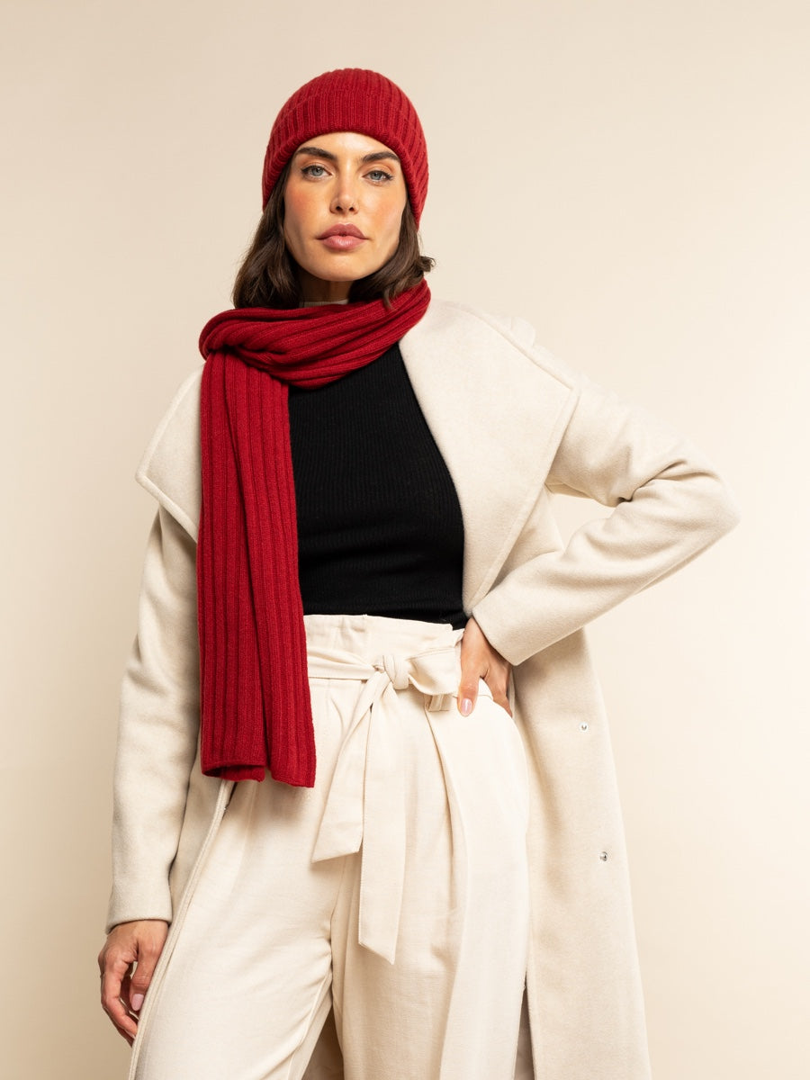 Woman wearing a red cashmere beanie and scarf with a white coat and pants on a beige background