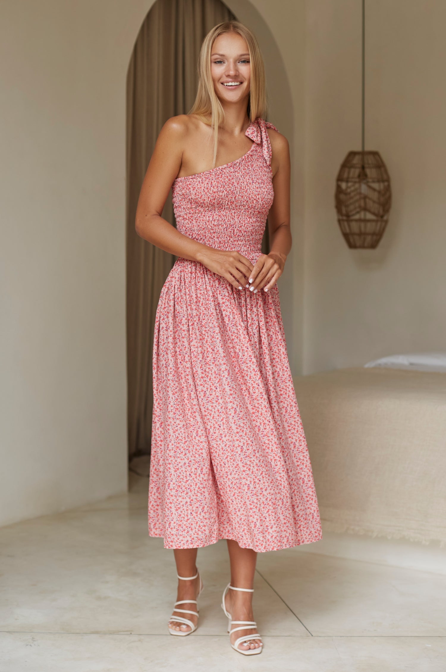 Woman wearing pink floral one-shoulder dress with a fitted bodice, smocked at the bust, midi length, front view