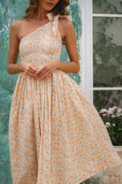Woman wearing a cream floral one-shoulder dress against a blurred outdoor background