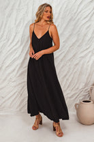 Woman wearing a black flowy maxi dress with deep v-neckline and spaghetti straps against a textured white wall. 