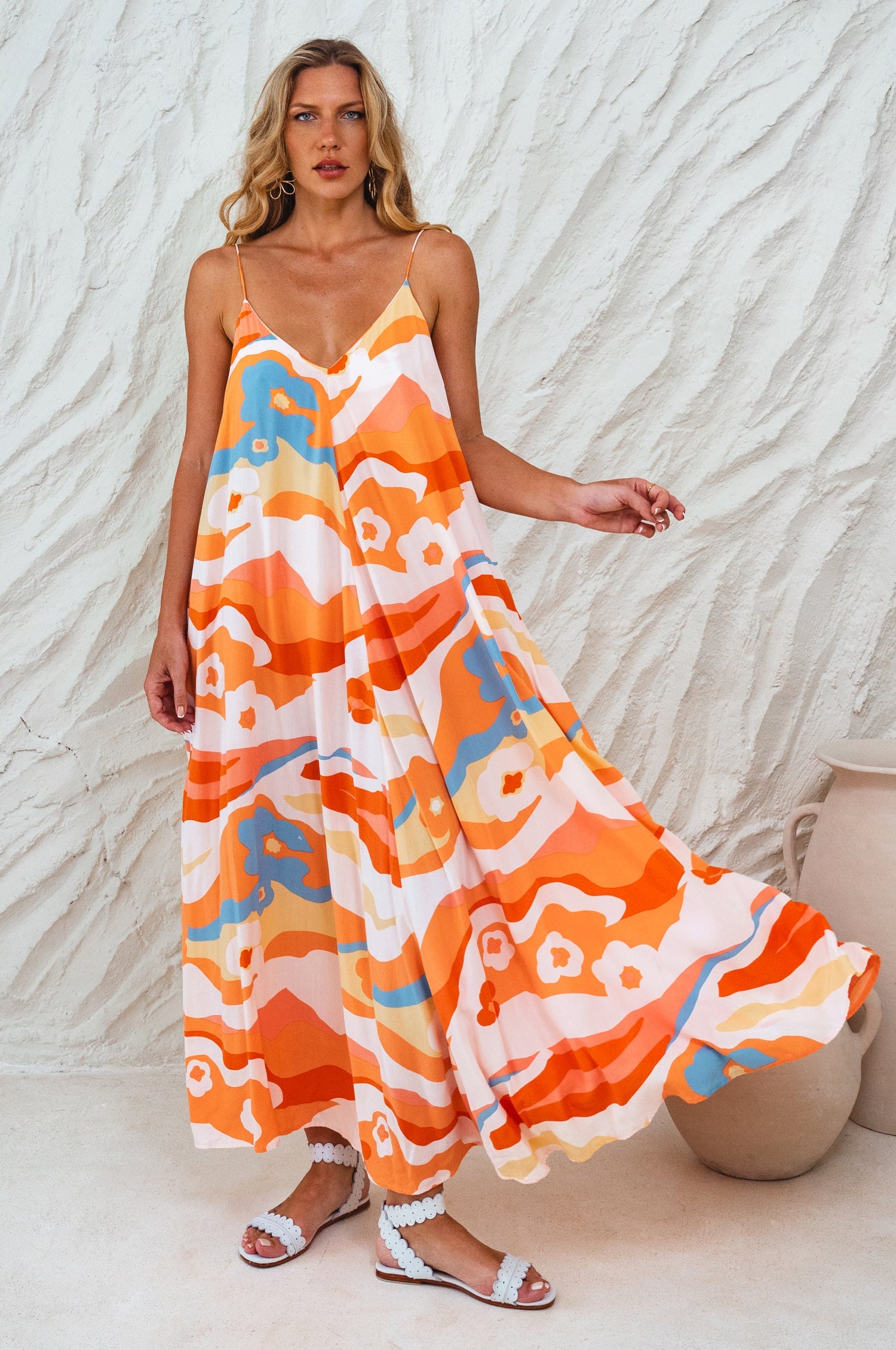 Woman wearing an orange colorful patterned flowy maxi dress with deep v-neckline and spaghetti straps against a white wall, front view against a textured white wall.