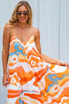Woman wearing an orange colorful abstract patterned flowy maxi dress with deep v-neckline and spaghetti straps against a light background