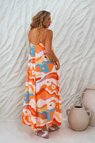 Woman wearing an orange colorful abstract patterned flowy maxi dress with deep v-neckline and spaghetti straps against a textured white wall.