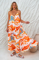 Woman wearing an orange colorful patterned flowy maxi dress with deep v-neckline and spaghetti straps against a white wall, front view against a textured white wall.