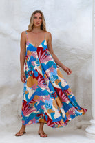 Woman wearing a colorful abstract floral flowy maxi dress with deep v-neckline and spaghetti straps against a white wall, front view.