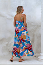 Woman wearing a blue colorful abstract floral flowy maxi dress with deep v-neckline and spaghetti straps against a white wall, front view against a white marble wall, back view