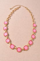 A gold-tone chain necklace featuring alternating pink cabochon stones and small gold spacer beads, displayed on a textured beige background.