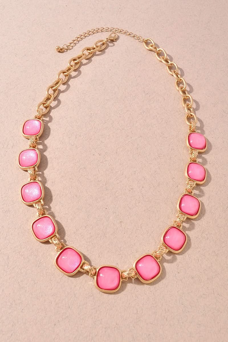 A gold-tone chain necklace featuring alternating pink cabochon stones and small gold spacer beads, displayed on a textured beige background.